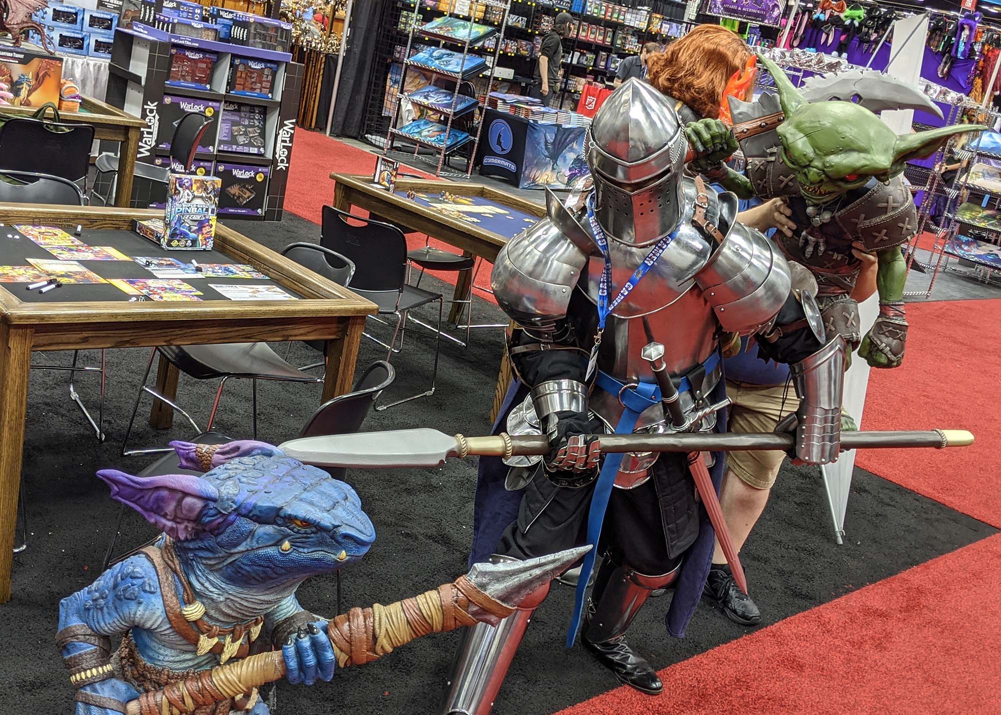 A person dressed in knight's armor points their spear at a blue foam kobold while a foam goblin stands at their back