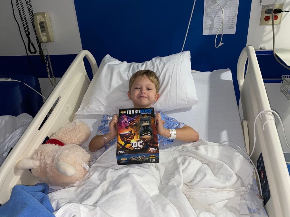 a photo of a smiling kid in a hospital bed, holding up a DC comics FunkoPop box