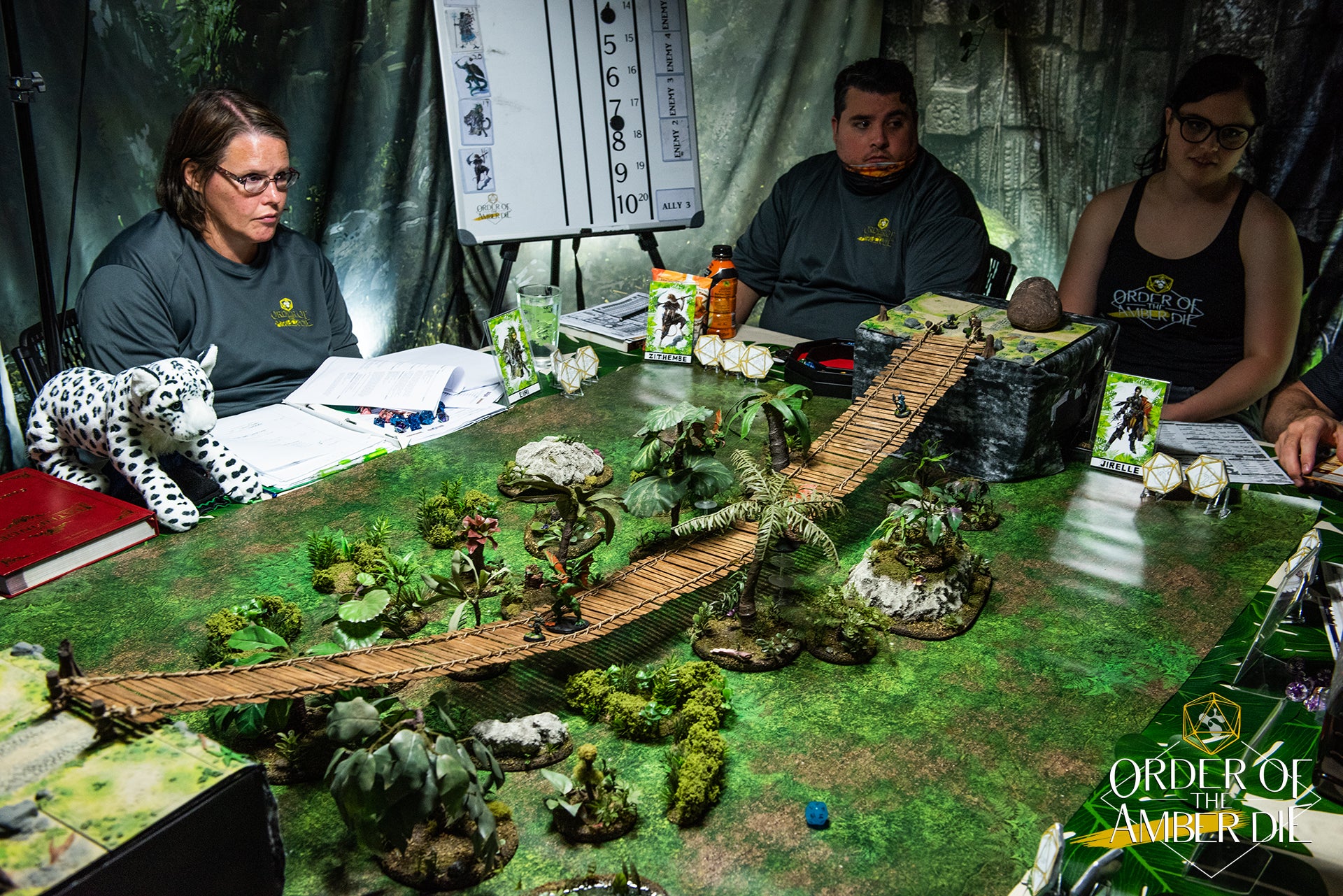 The order of the Amber Die players sit around a table decorated with mini figures and landscaping to represent a jungle