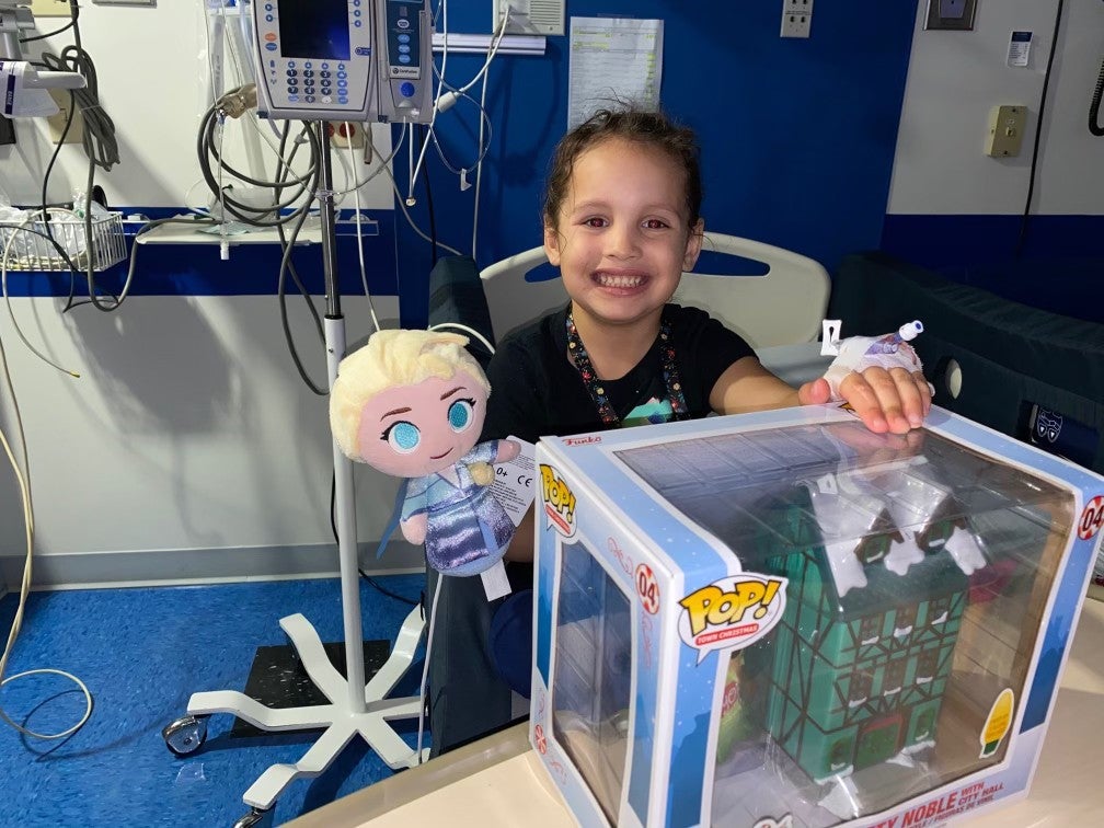 a photo of a smiling kid in a hospital room with an Elsa doll and Funko Pop figure