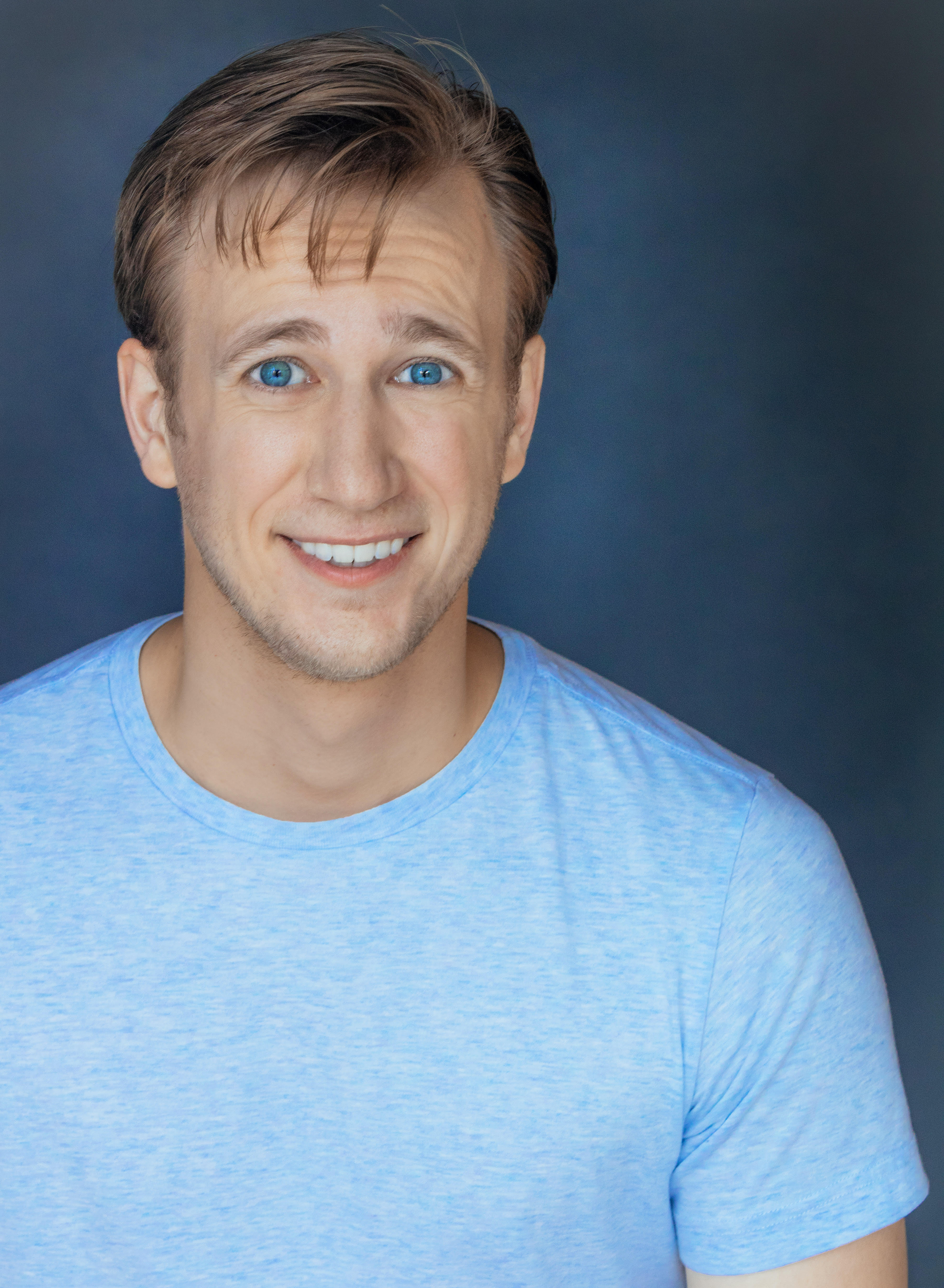 Headshot of Nathan Ondracek, smiling in front of a dark blue background while wearing a light blue t-shirt