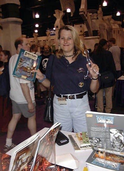 Lisa Stevens standing in a convention booth, holding up a book