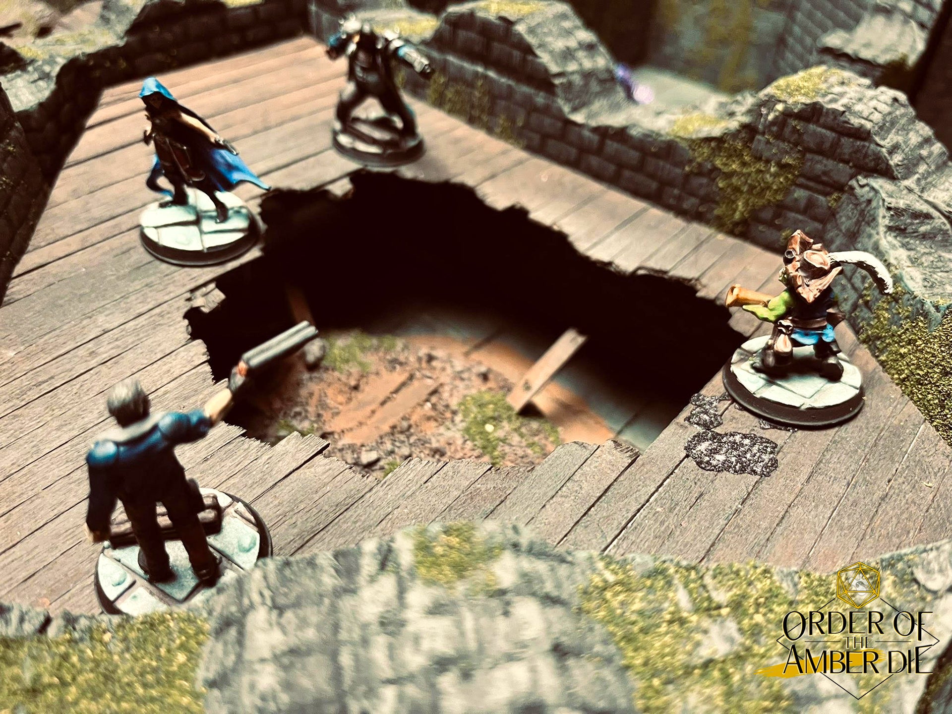 Order of the Amber Die: A top down view of four mini figures standing on a wooden platform looking into a hole that leads deeper into the ruins they're investigating