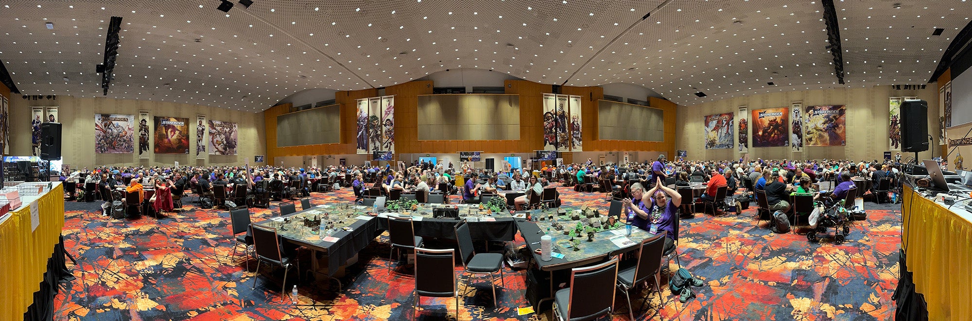 Gen Con 2025 Sagamore Ballroom: A panoramic photo of a ballroom full of tables packed with ttrpg players, at the bottom center of the photo are three tables covered in miniatures and terrain shaped in a U formation. Two game masters are sitting at the tables