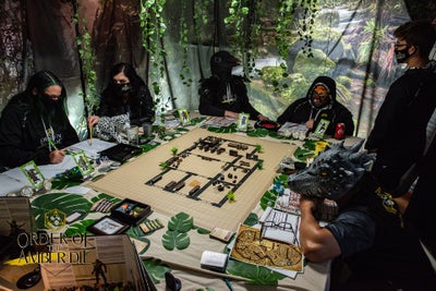 The order of the Amber Die, all wearing masks, sitting around the gaming table