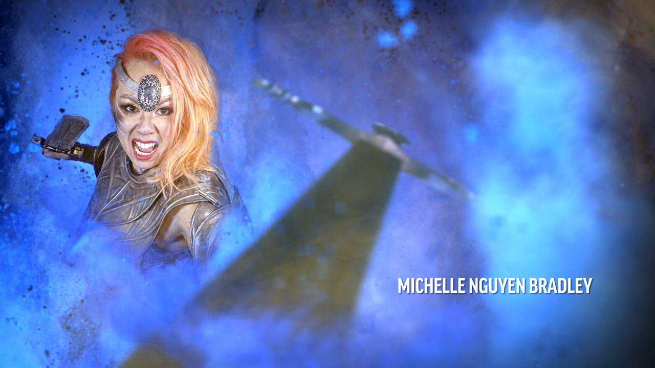 Michelle Nguyen Bradley, a woman in armor stands with a sword raised in a blue smokey background with a sword pointed at the viewer in the foreground