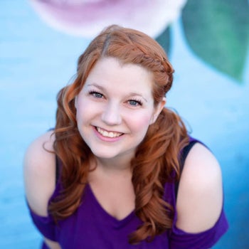 Headshot of Bonnie Gordon. Her hair is braided back from her face and she's wearing a purple off shoulder blouse, standing in front of a blue background