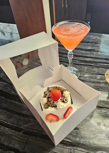 A picture of a Strawberry Machine Cake, a small cake decorated with white frosty, chocolate cogs, and strawberries. Behind the cake is a triangular cocktail glass with a bright pink cosmopolitan drink.