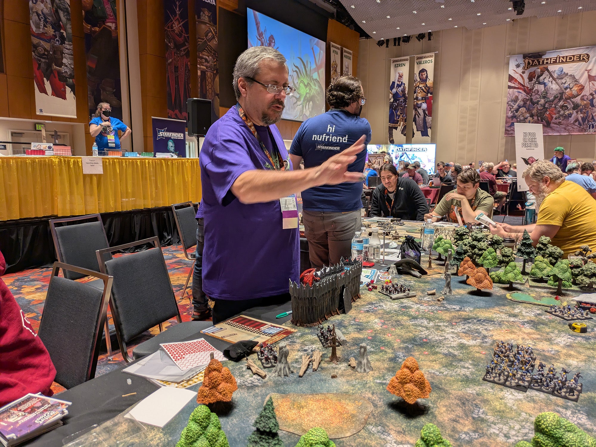 A photo of a GM (a game master) in a purple shirt gesturing over a table covered in miniatures and terrain as he guides players (off camera) through a campaign