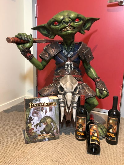 life sized goblin statue sitting next two the pathfinder bestiary and three bottles of wine