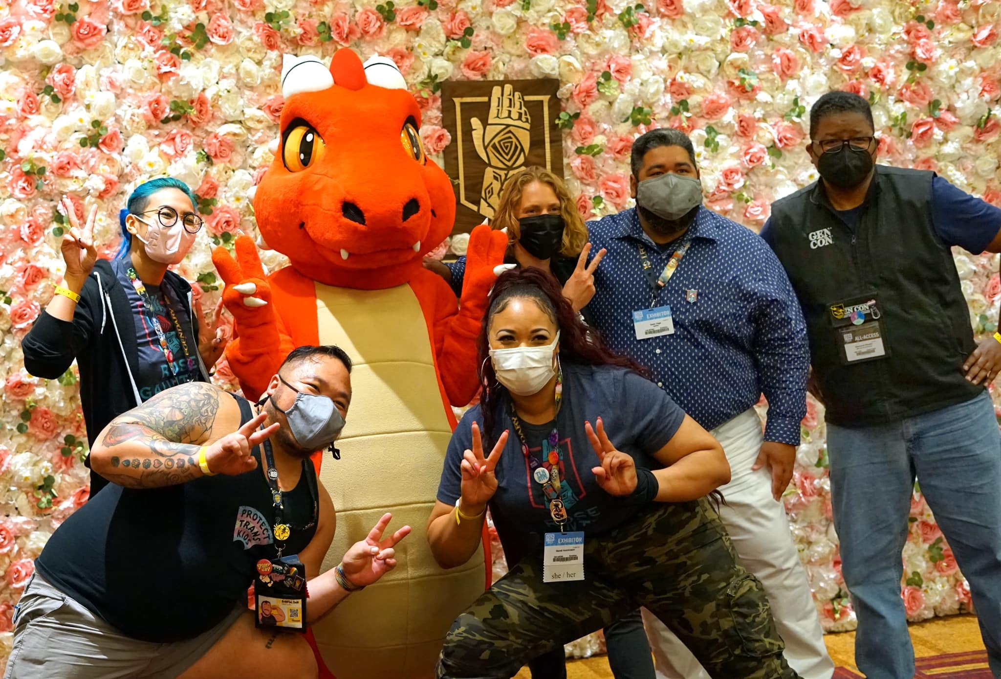 A group of BIPOC gamers and Genevieve the Dragon standing in front of a wall of roses.
