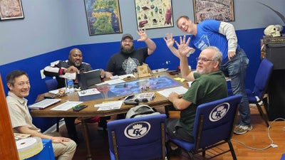 A group of five tabletop gamers sitting at an octagonal table covered in maps, miniatures, and character sheets.