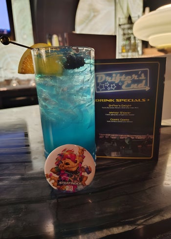 A photo of a bright blue cocktail in a tall glass with a button of a skittermander in front of it and a propped up Drifter's End Cocktail menu in the background