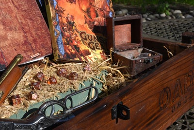 A staged photo of Pathfinder Guns and Gears in a wooden cheset sitting on a bed of hay with amber colored dice and other small adventuring accessories.