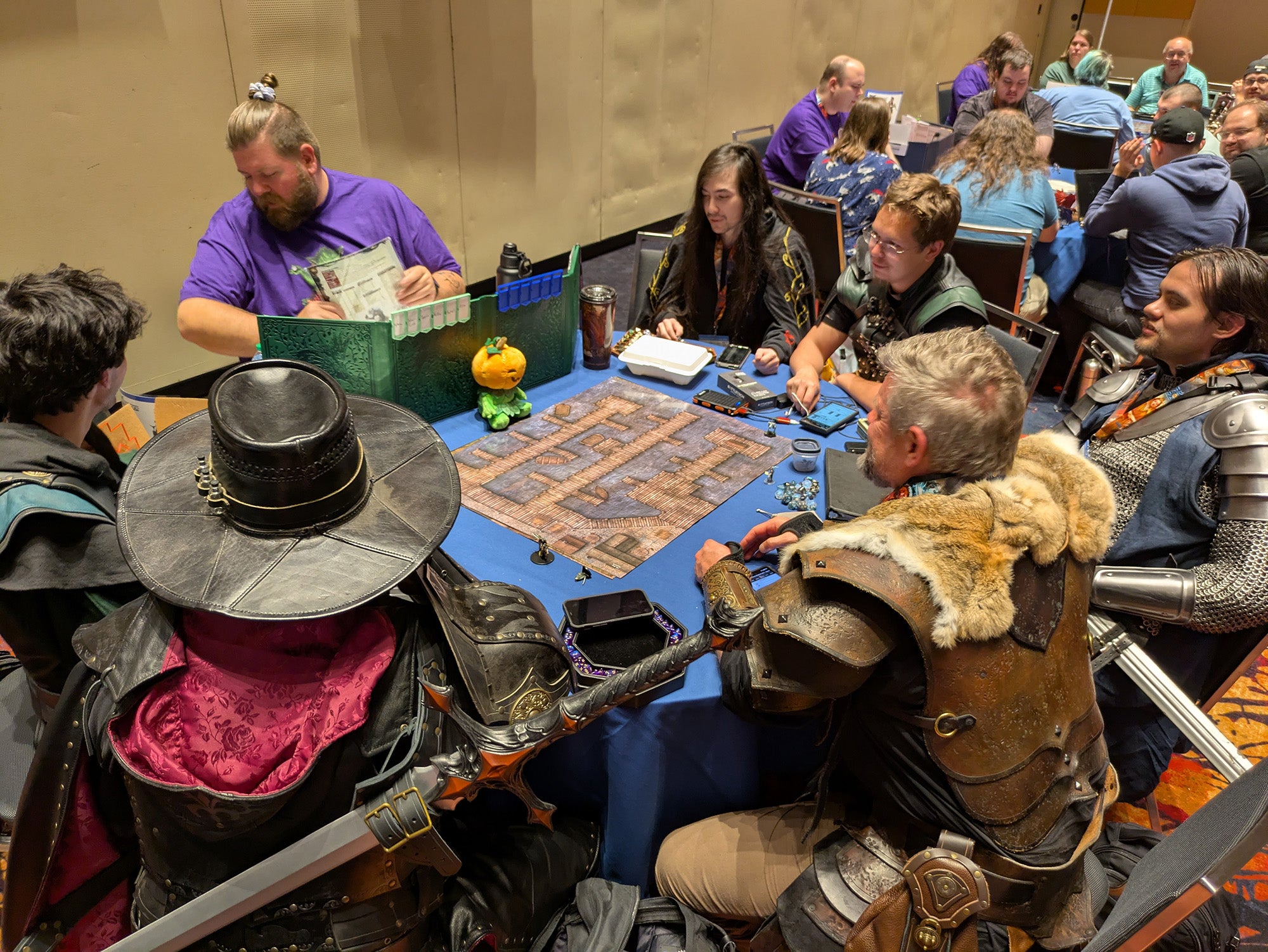 A photo of a table full of pathfinder society players. The game master is wearing a purple t-shirt while all the players are dressed in full chainmail and leather armor