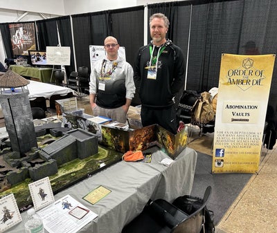 Two men standing in a convention center booth next to a table covered in miniature terrain and buildings