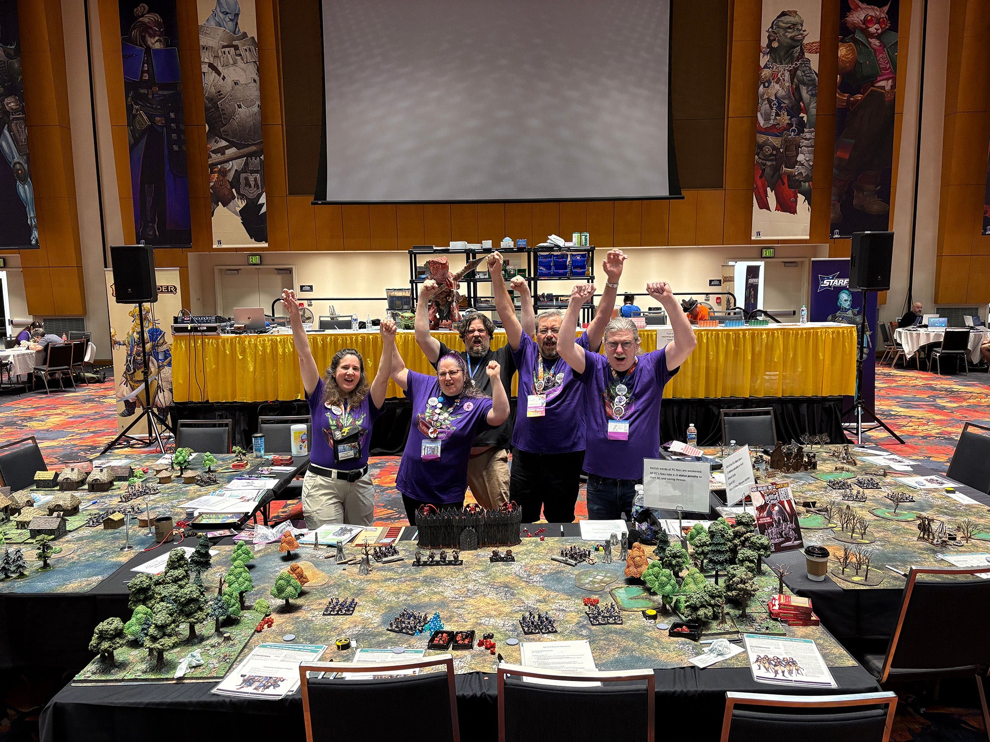 A photo of a group of five people wearing purple t-shirts standing and cheering as they're surrounded by tables covered in miniatures on three sides