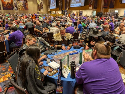 Gen Con 2025 Sagamore Ballroom:  photo of a ballroom full of tables packed with ttrpg players. The photo is centered on a table at the bottom of the image full of players dressed in full fantasy armor