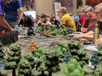 Gen Con 2025 Sagamore Ballroom: A close up shot of a table covered in a miniatures creating the scene of a fortress in a forest under siege