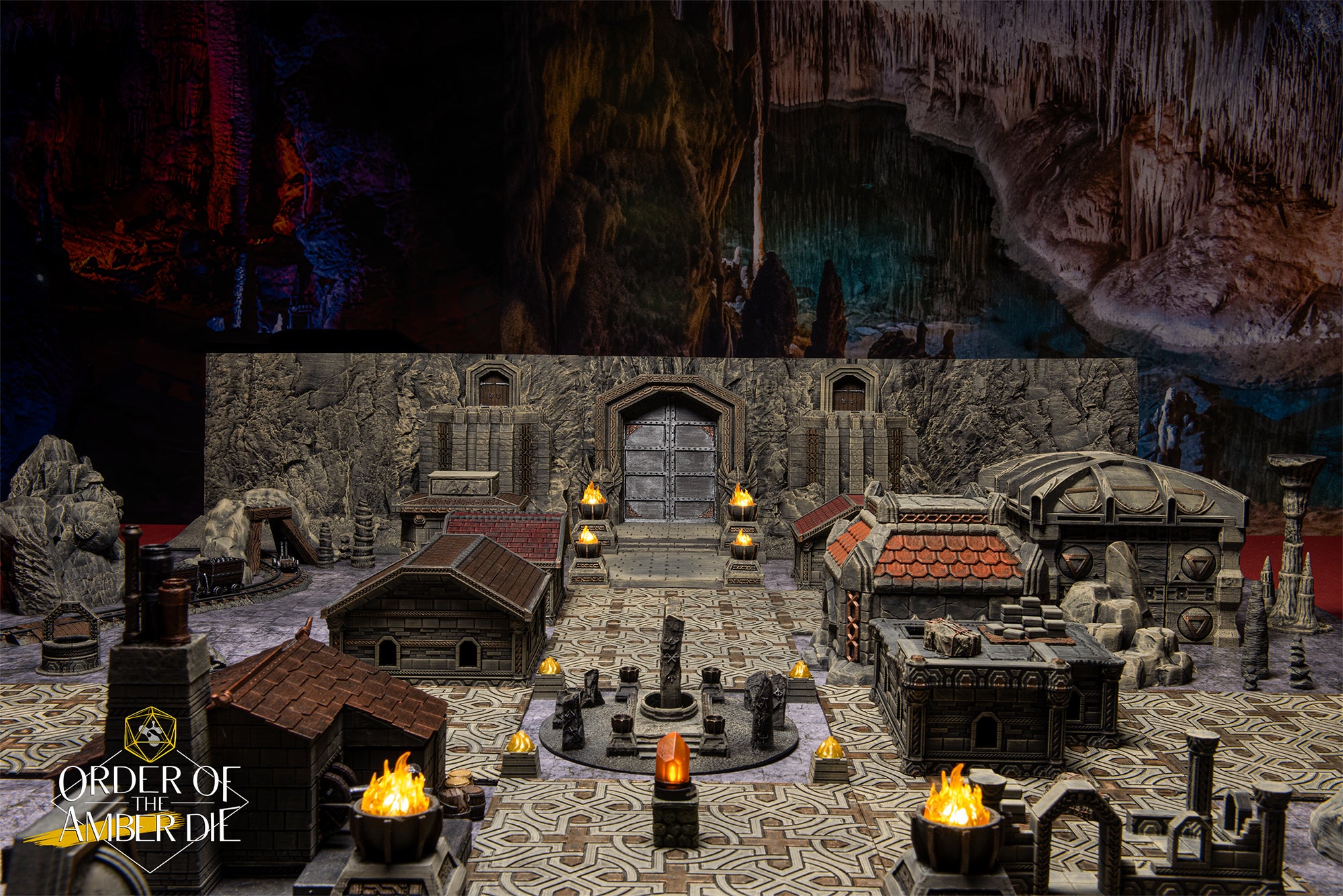 A table with a model down setup featuring miniature stone buildings, towers, flaming lanterns, and a stone statue in the center of town