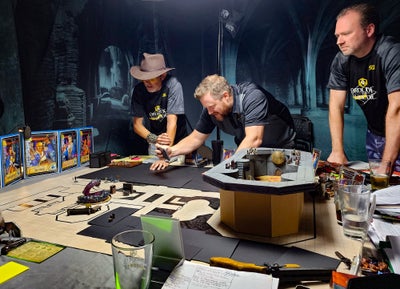 The abomination arsenal, Order of the Amber Die: three men in matching black t-shirts lean over a table covered in game maps, miniatures, water glasses, and character sheets.