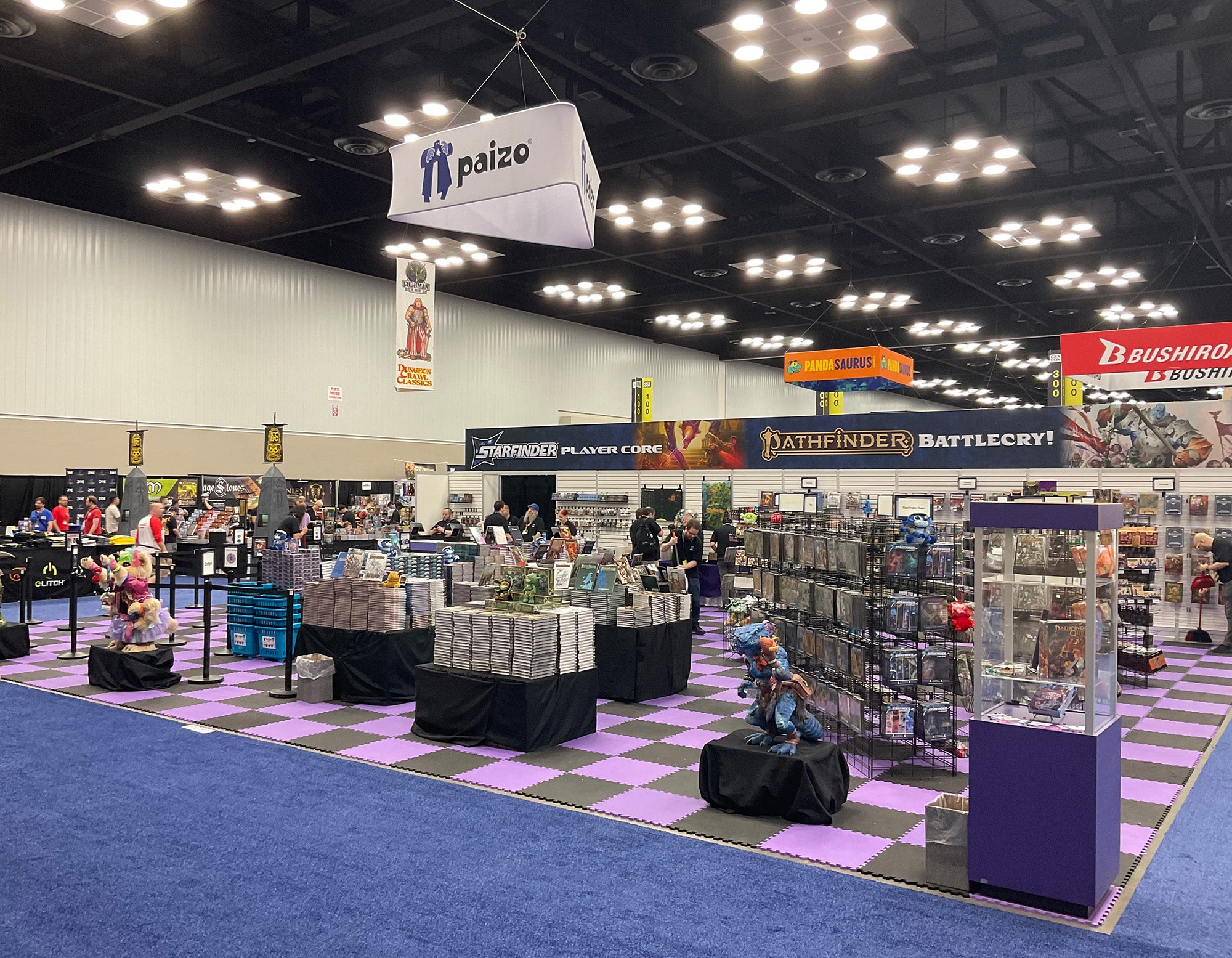 A photo of Paizo's booth at GenCon 2025 before the show floor opens with it's iconic purple and black checkered floor mat. The show floor and booth are empty of people, but full of product.