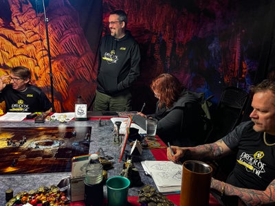 The Order of the Amber Die tabletop gaming group sitting around a gaming table with a large map and props in the center of the table. One man is standing at the corner of the table smiling at someone off camera